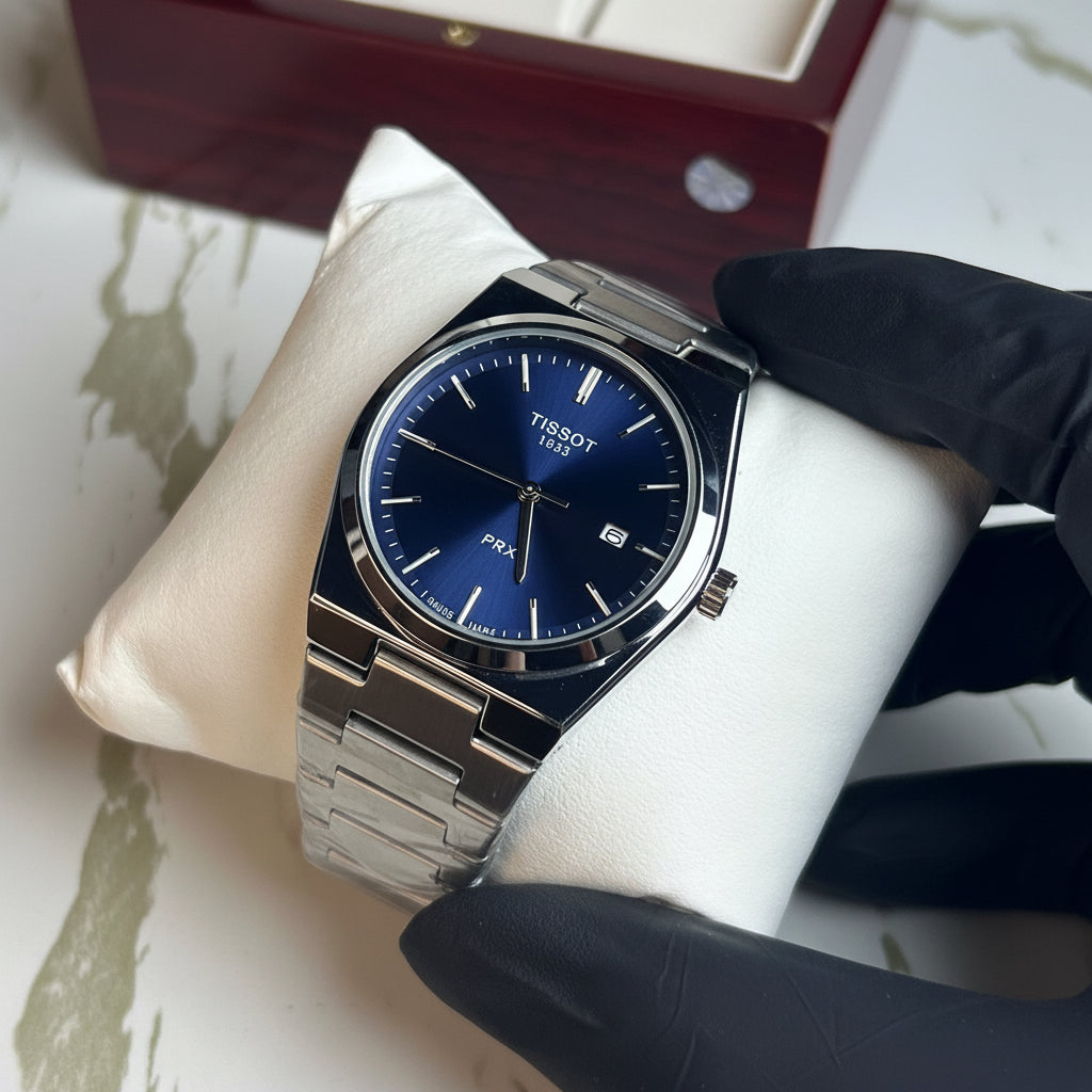 A Tissot PRX watch with a blue dial and stainless steel case, displayed on a white cushion held by a hand wearing a black glove, with a wooden presentation box in the background.
