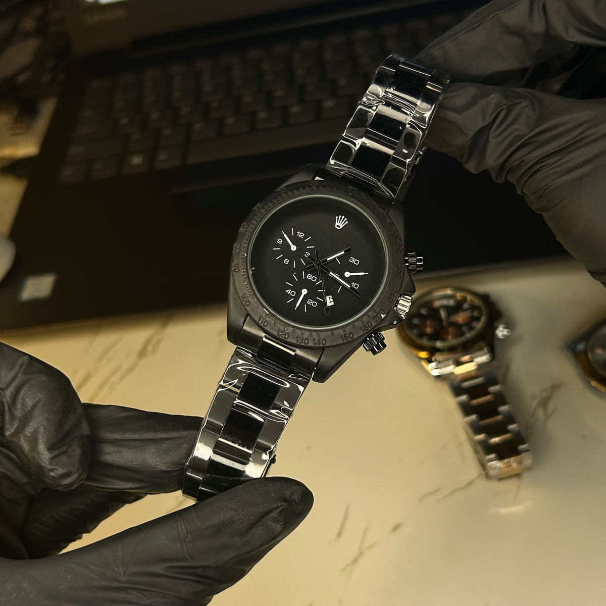 A black stainless steel chronograph watch with a black dial and silver-tone links, held by hands wearing black gloves. The Rolex crown logo is visible on the watch face, and a second watch is blurred in the background on a light marble surface.