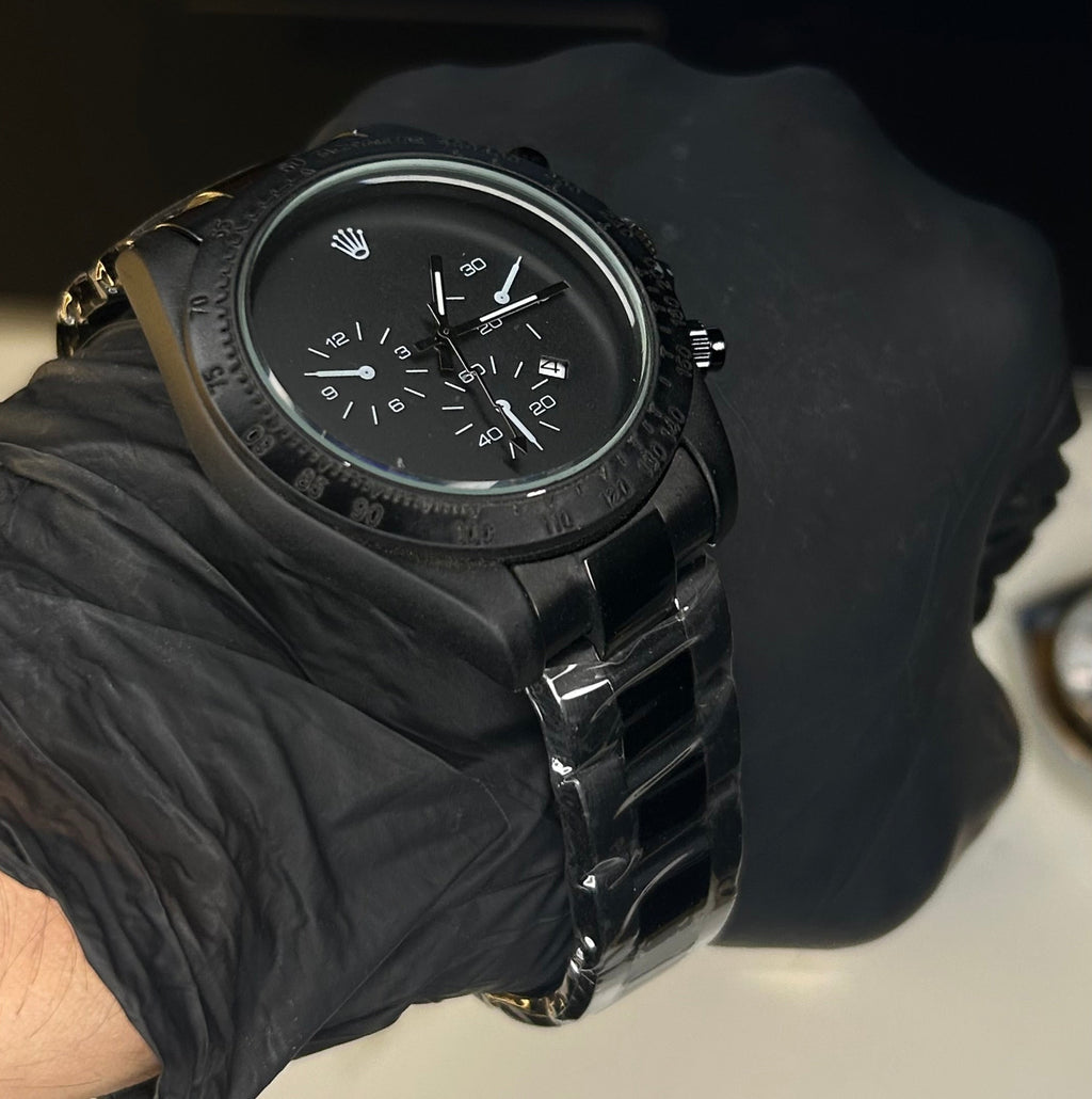 A black chronograph watch with a black metal link bracelet, worn on a wrist covered in a black glove. The Rolex crown logo is visible on the dial, and the background shows a blurred laptop keyboard.