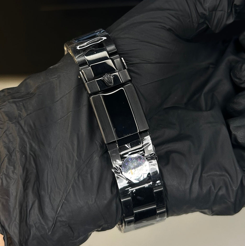 A close-up of a shiny black metal link bracelet being held on a wrist by a hand wearing a black glove, with a blurred laptop keyboard in the background.