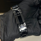 A close-up of a shiny black metal link bracelet being held on a wrist by a hand wearing a black glove, with a blurred laptop keyboard in the background.