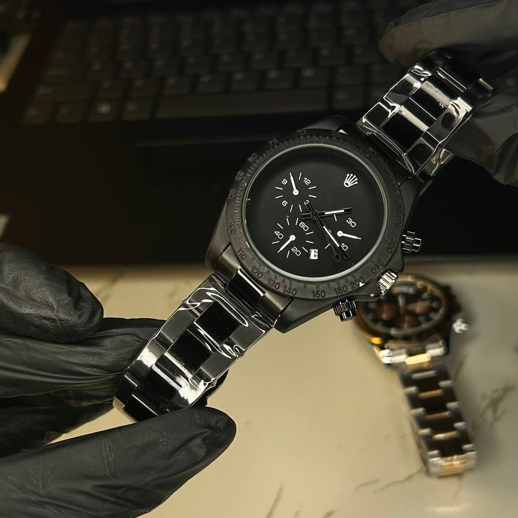 A black analog watch with a black dial and a silver-toned metal link bracelet is held by hands wearing black gloves. The background shows a blurred laptop screen and a marble surface with another watch lying on it.