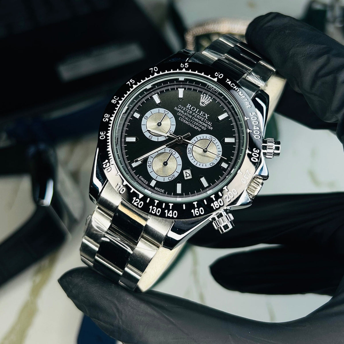 A stainless steel Daytona watch with a black dial and silver-tone bracelet, held by a hand wearing a black glove. The watch is positioned in front of a blurred laptop and desk background.