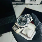 A stainless steel wristwatch with a black dial and Roman numeral markers is held by a hand wearing a black glove. The watch has a fluted bezel and a multi-link bracelet.