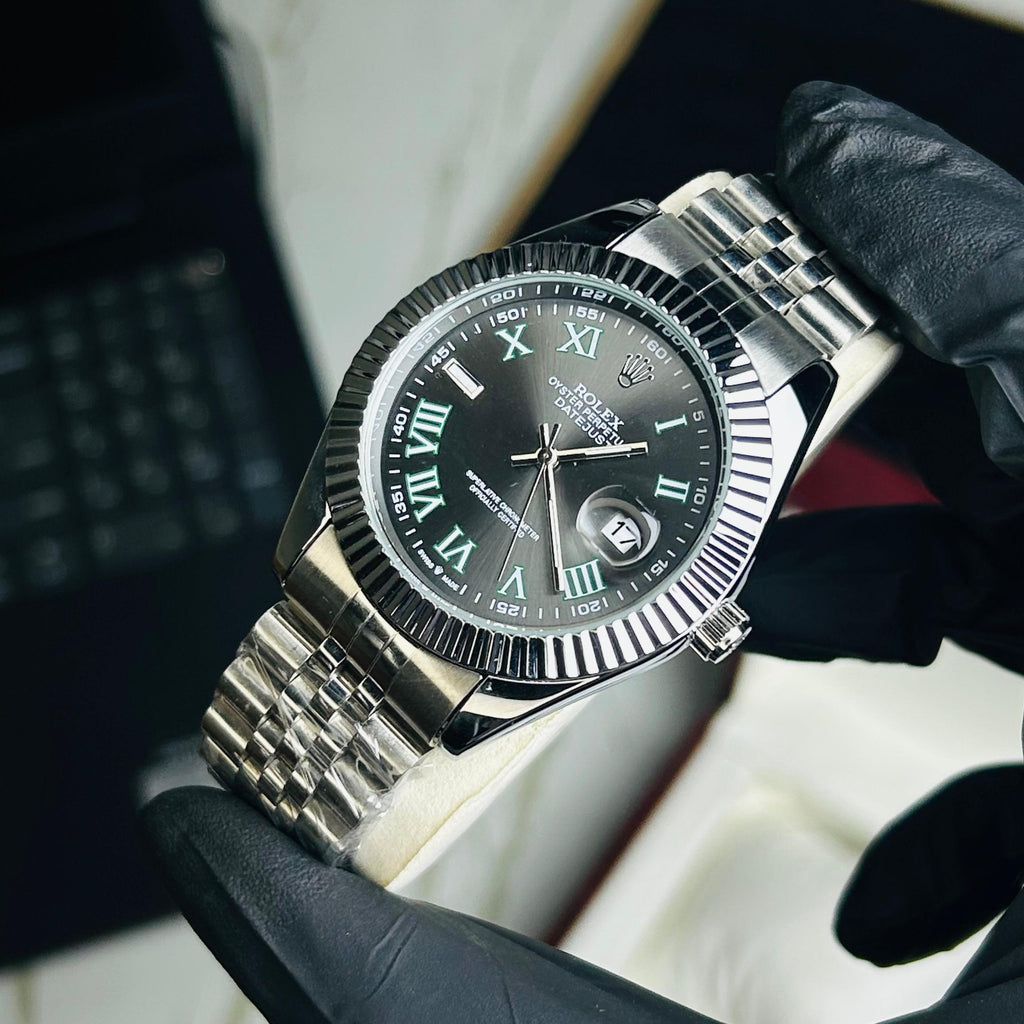 A stainless steel wristwatch with a black dial and Roman numeral markers is held by a hand wearing a black glove. The watch has a fluted bezel and a multi-link metal bracelet.