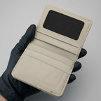 A close-up of a light beige leather wallet held by a hand wearing a black glove. The wallet is open, showing a black mesh ID window on the inside cover, multiple card slots, and a textured leather interior.