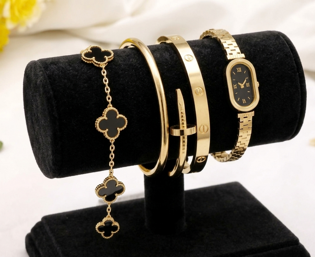 A gold-tone jewelry display stand holding a bracelet with four clover motifs and several gold bangles and a watch with black faces, set against a soft white background with a blurred yellow flower.