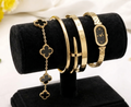 A gold-tone jewelry display stand holding a bracelet with four clover motifs and several gold bangles and a watch with black faces, set against a soft white background with a blurred yellow flower.