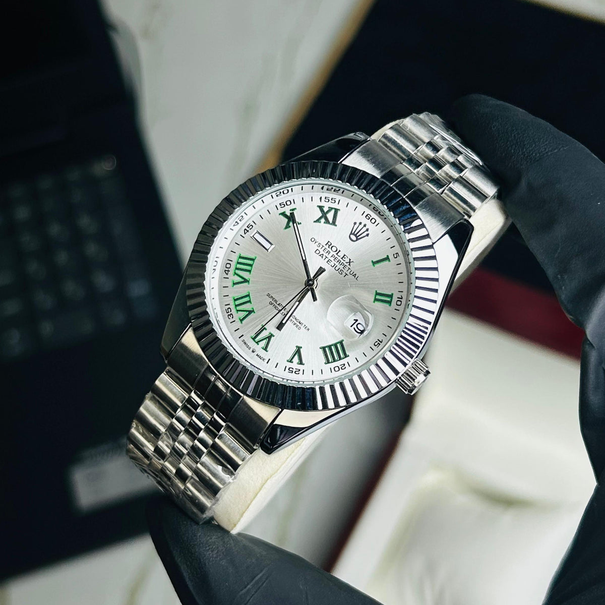 A Rolex Submariner Date watch with a silver-tone metal bracelet and a white dial featuring green Roman numerals, held by a hand wearing a black glove.